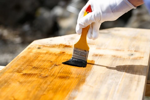 Main ouvrier appliquant un produit de finition sur planches de bois brut avec pinceau plat, portant gants de protection blancs