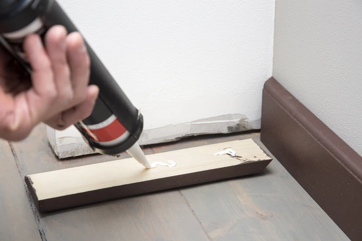 Intérieur d'atelier, artisan appliquant de la colle blanche avec un pistolet à colle sur une planche de bois claire posée sur établi