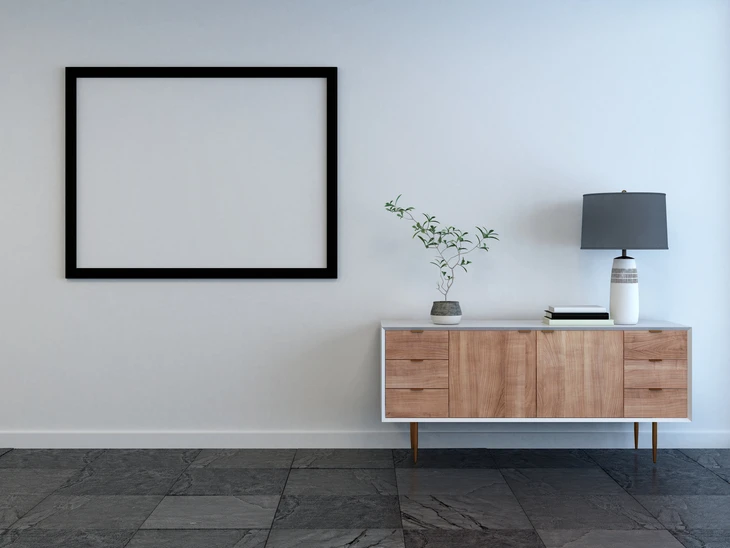 Intérieur moderne minimaliste avec meuble bas en bois naturel sur pieds effilés, lampe contemporaine à abat-jour anthracite, petit arbre en pot et cadre noir vide au mur