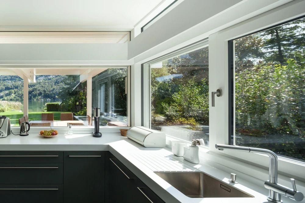 Cuisine moderne avec plan de travail blanc, façade en bois sombre et fenêtres panoramiques donnant sur jardin paysager