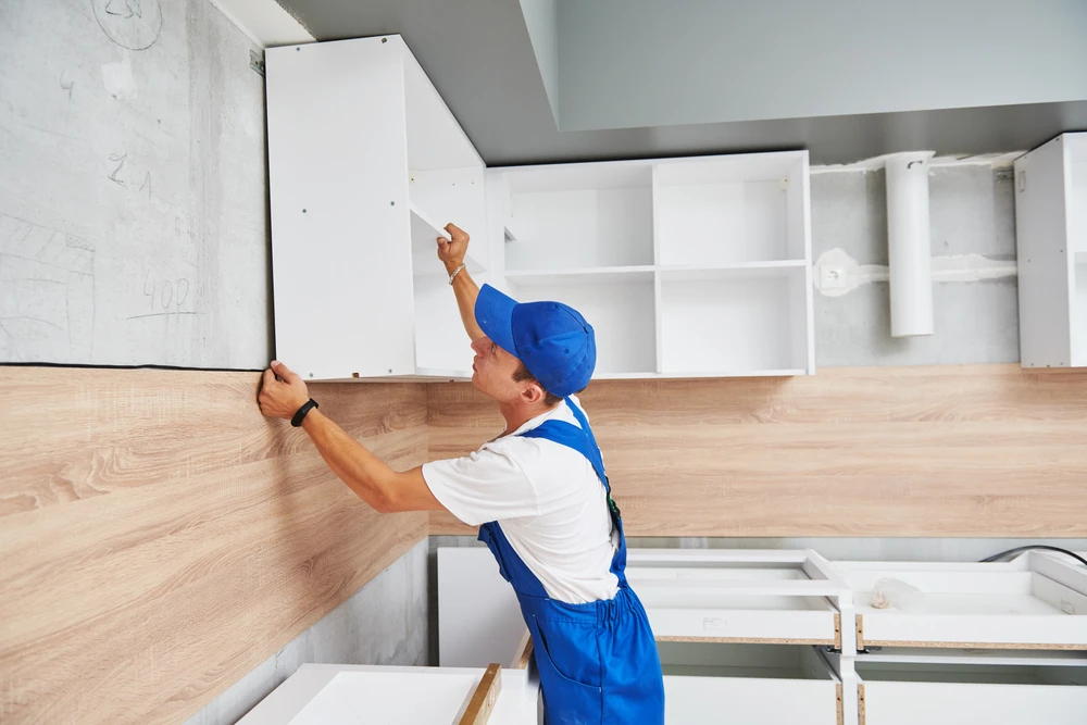 Un artisan en tenue de travail bleue et blanche avec casque installe une crédence en bois dans une cuisine moderne