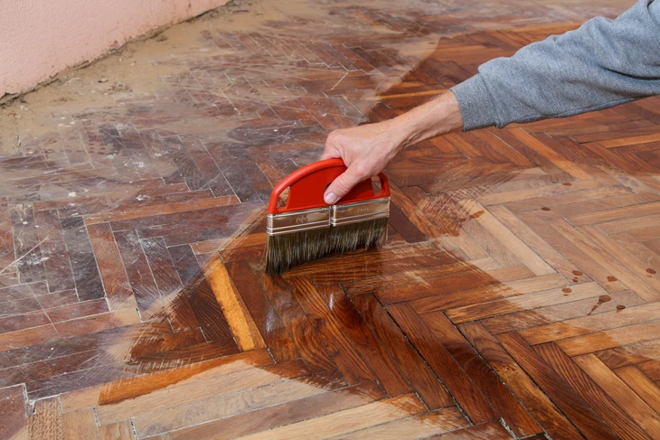 Une personne applique un produit de finition sur un parquet en bois massif à motif point de Hongrie avec un pinceau plat à manche rouge