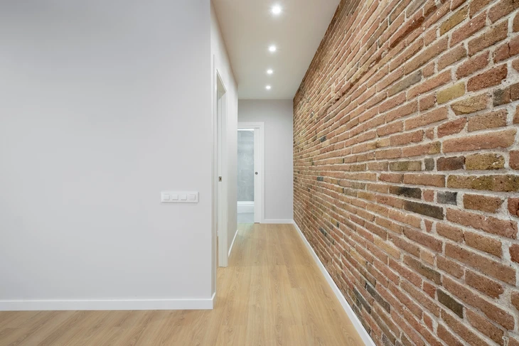 Un couloir moderne bien éclairé avec un plancher en bois élégant et un mur en briques apparentes, menant à une porte lointaine