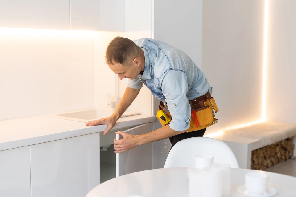Un artisan en chemise en jean installe des éléments de cuisine blanche moderne dans un intérieur contemporain aux murs blancs