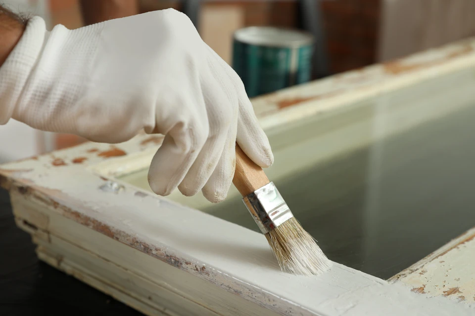 Intérieur d'atelier, personne portant gants blancs appliquant peinture au pinceau sur meuble en bois clair