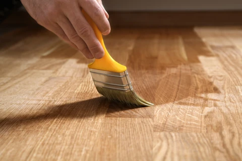 Une main tenant un pinceau plat à manche orange applique un produit de finition sur un parquet en bois naturel aux veines apparentes