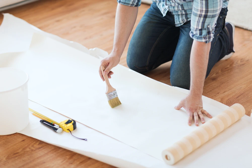 Une personne en chemise à carreaux applique une finition au pinceau sur un panneau de bois blanc posé au sol dans un intérieur aux parquets clairs