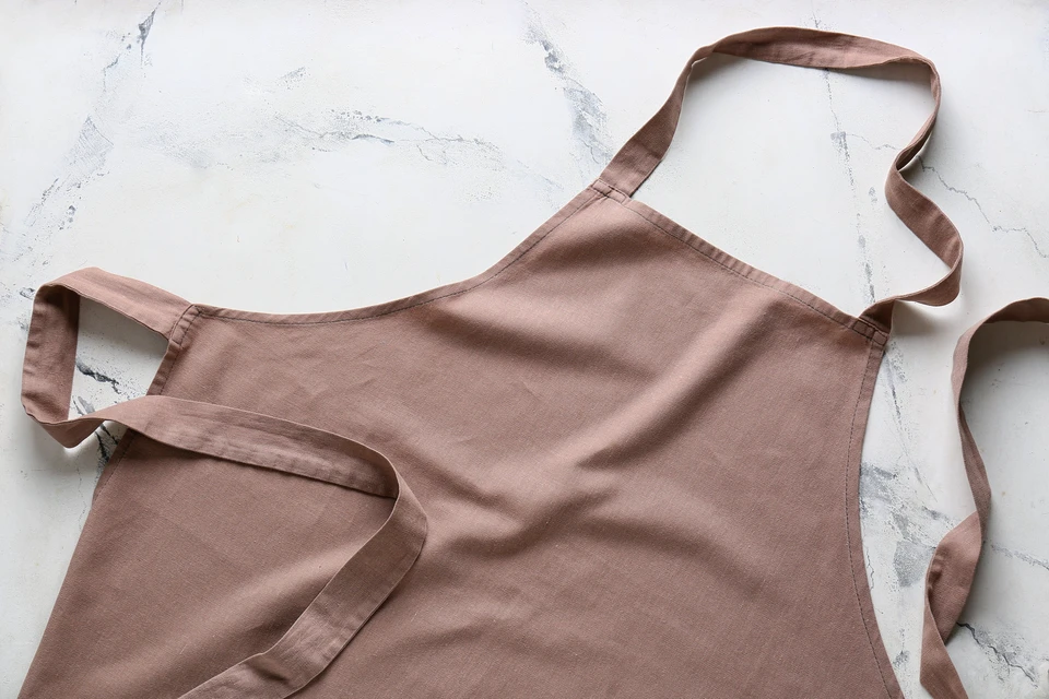 Tablier de cuisine en lin beige disposé à plat sur surface marbrée blanche