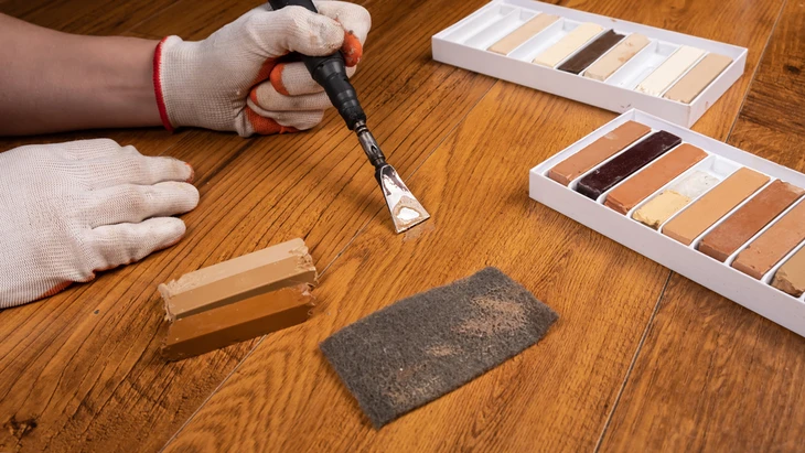 Une personne portant des gants blancs applique un produit de réparation sur du parquet en bois avec un outil de précision