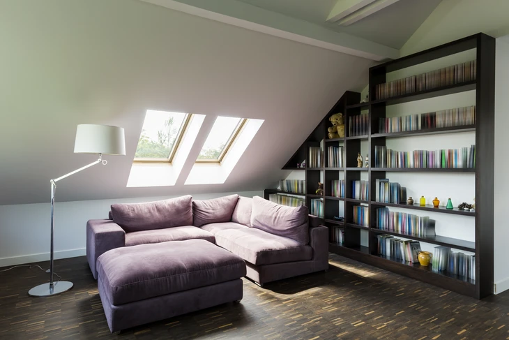 Salon moderne sous combles avec canapé d'angle violet et repose-pieds, bibliothèque noire intégrée sous pente, lampadaire design blanc